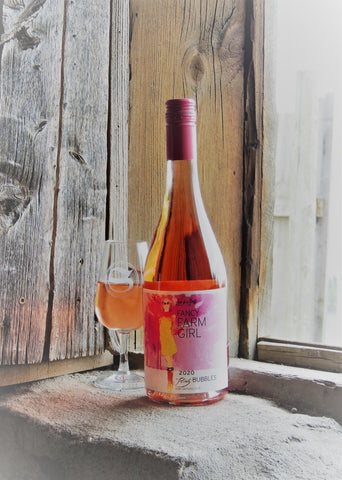 A photo of a glass and bottle of Fancy Farm Girl Flirty Bubbles sparkling rosé in the window of a barn, lit by natural sunlight